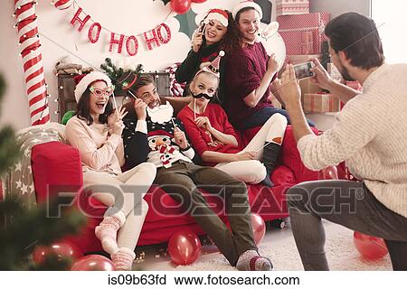 Stock Image - Young man photographing friends on sofa at christmas party. Fotosearch