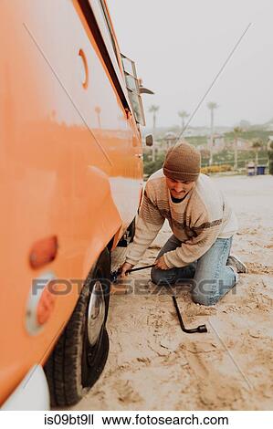 Stock Image - Young man removing flat tyre on recreational vehicle at beach, Jalama, California, USA. Fotosearch