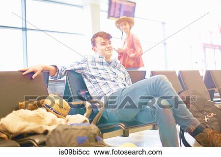Stock Image - Young man sitting at airport, backpack beside him, young woman standing nearby, looking towards him. Fotosearch
