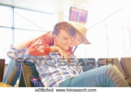Stock Image - Young man sitting at airport, young woman reaching over seat to hug him. Fotosearch