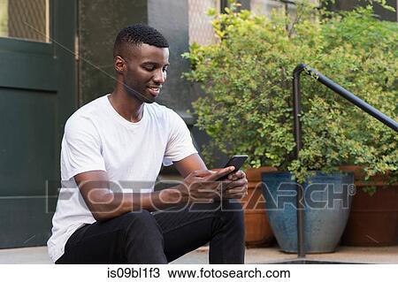 Stock Image - Young man using cellphone on step in streets of New York, US. Fotosearch