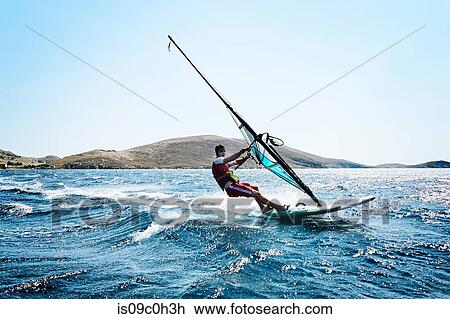 Stock Image - Young man windsurfing ocean waves, side view, Limnos, Khios, Greece. Fotosearch