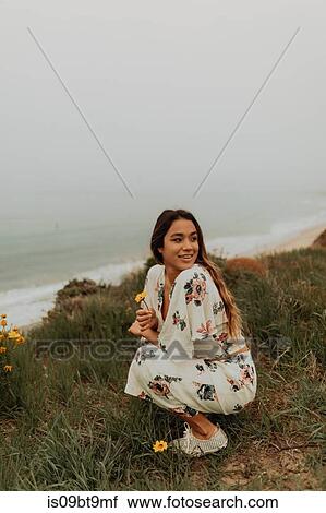 Stock Image - Young woman crouching and holding wildflowers at coast, Jalama, California, USA. Fotosearch