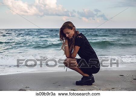 Stock Image - Young woman crouching by water's edge on beach, Odessa, Odessa Oblast, Ukraine. Fotosearch