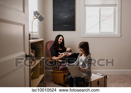 Young woman holding baby son in living room armchair while daughter reads book View Large Photo Image Picture - Young woman holding baby son in living room armchair while daughter reads book. Fotosearch