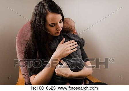 Stock Image - Young woman holding baby son in living room armchair. Fotosearch