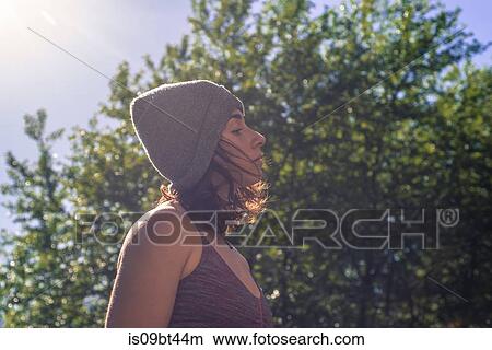 Picture - Young woman in knitted hat contemplating in front of tree foliage, backlit head and shoulder 0ofile. Fotosearch