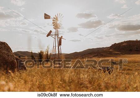 Stock Image - Young woman in long dress standing on traditional wind turbine, Exeter, California, USA. Fotosearch