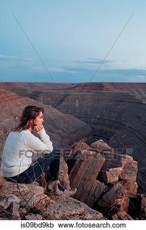 Stock Image - Young woman in remote setting, sitting on rocks, looking at view, Mexican Hat, Utah, USA. Fotosearch