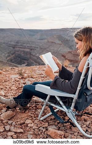 Stock Photograph - Young woman in remote setting, sitting on camping chair, reading book, Mexican Hat, Utah, USA. Fotosearch