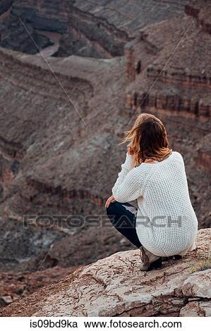 Stock Image - Young woman in remote setting, crouching on rocks, looking at view, rear view, Mexican Hat, Utah, USA. Fotosearch