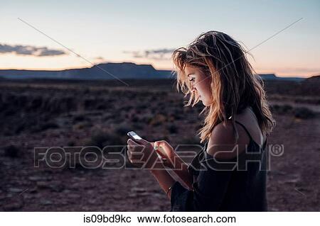 Stock Image - Young woman in remote setting, using smartphone, Mexican Hat, Utah, USA. Fotosearch
