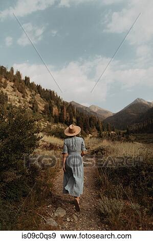 Stock Photo - Young woman in stetson and maxi dress strolling on rural valley dirt track, rear view, Mineral King, California, USA. Fotosearch