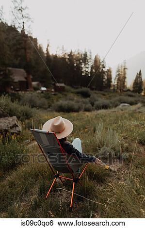 Stock Photograph - Young woman in stetson relaxing on deck chair in rural valley, rear view, Mineral King, California, USA. Fotosearch