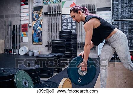 Stock Photograph - Young woman lifting weight plate in gym. Fotosearch