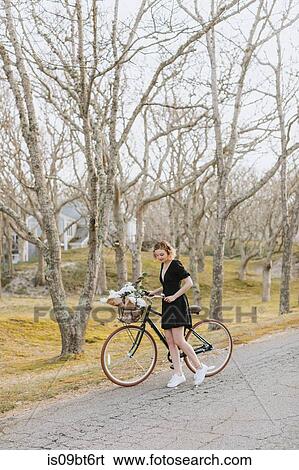 Stock Photograph - Young woman pushing bicycle with bunch of flowers on rural road, Menemsha, Martha's Vineyard, Massachusetts, USA. Fotosearch