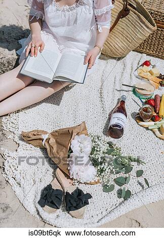 Stock Image - Young woman reading book on beach, mid section, Menemsha, Martha's Vineyard, Massachusetts, USA. Fotosearch