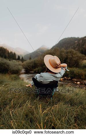 Stock Image - Young woman relaxing in deckchair by rural river, rear view, Mineral King, California, USA. Fotosearch
