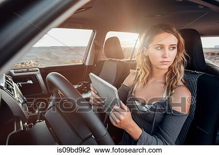Stock Image - Young woman sitting in vehicle, holding digital tablet, looking out of window, Mexican Hat, Utah, USA. Fotosearch