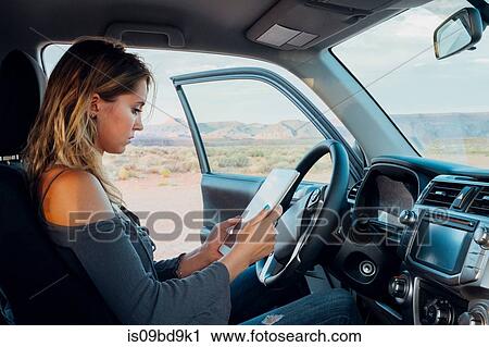 Stock Image - Young woman sitting in vehicle, looking at digital tablet, Mexican Hat, Utah, USA. Fotosearch