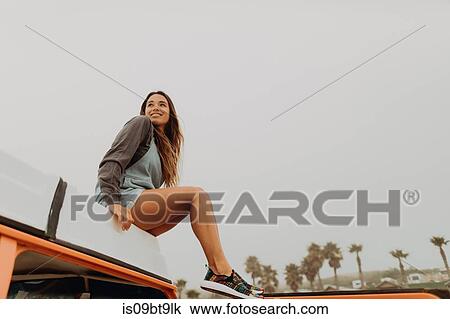 Stock Image - Young woman sitting on recreational vehicle roof at beach, Jalama, California, USA. Fotosearch
