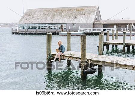 Stock Image - Young woman sitting on wooden pier, portrait, Menemsha, Martha's Vineyard, Massachusetts, USA. Fotosearch