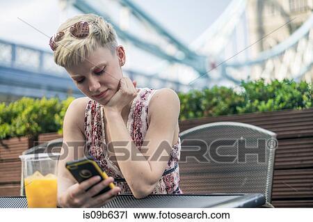 Stock Image - Young woman sitting outdoors, plastic drinking glass in front of her, looking at smartphone. Fotosearch