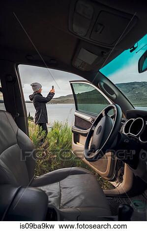 Stock Photo - Young woman standing beside Dillon Reservoir, holding smartphone, view through parked car, Silverthorne, Colorado, USA. Fotosearch