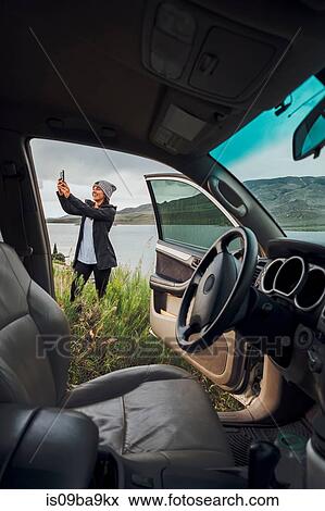 Stock Photo - Young woman standing beside Dillon Reservoir, holding smartphone, view through parked car, Silverthorne, Colorado, USA. Fotosearch