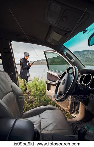 Stock Photograph - Young woman standing beside Dillon Reservoir, holding smartphone, view through parked car, Silverthorne, Colorado, USA. Fotosearch