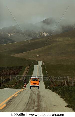 Stock Image - Young woman walking toward recreational vehicle on rural road, Jalama, California, USA. Fotosearch