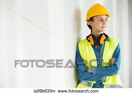 Young woman, wearing hi vis vest and hard hat, leaning against wall, arms folded View Large Photo Image Picture - Young woman, wearing hi vis vest and hard hat, leaning against wall, arms folded. Fotosearch