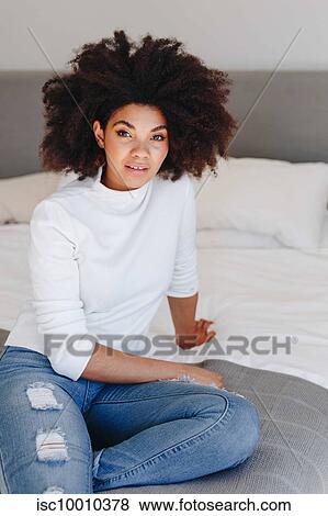 Stock Photo - Young woman with afro hairstyle sitting on bed, portrait. Fotosearch