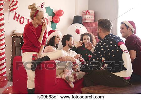 Stock Image - Young women and men eating popcorn on sofa at christmas party. Fotosearch