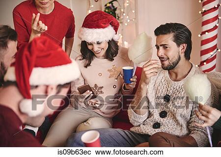 Stock Image - Young women and men looking at smartphone on sofa at christmas party. Fotosearch