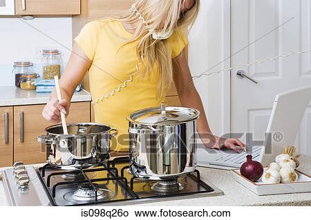 Picture - Woman cooking whilst on telephone and using laptop. Fotosearch