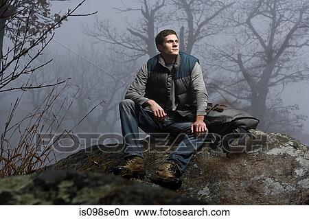 Picture - A male hiker sitting on a rock in a misty forest. Fotosearch