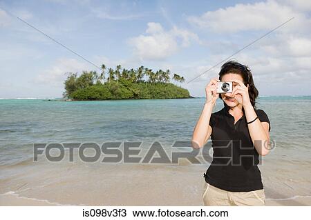 Stock Image - Woman taking photograph on beach with island in background. Fotosearch