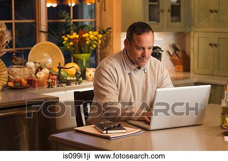 Stock Image - Mature man working on laptop in kitchen. Fotosearch