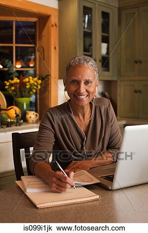 Stock Image - Mature woman using laptop and wearing ear phones, portrait. Fotosearch