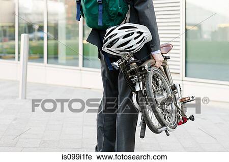 Stock Image - Mid adult businessman carrying folding bicycle. Fotosearch