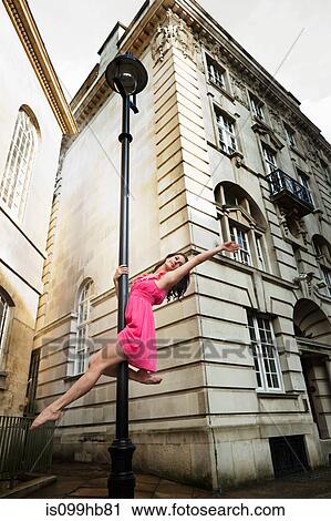 Stock Image - Mid adult woman in pink dress performing dance on street lamp in city. Fotosearch