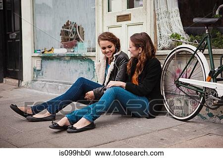 Stock Image - Two young women sitting on pavement outside cafe. Fotosearch