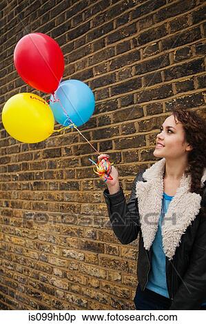 Stock Photography - Young woman holding colourful balloons against brick wall. Fotosearch