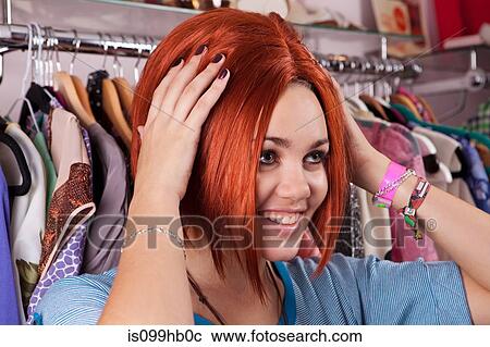 Stock Image - Young woman trying on wig in clothes shop, smiling. Fotosearch