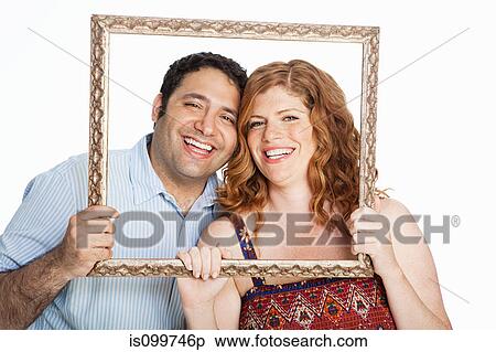 Stock Photography - Couple smiling throrugh picture frame against white background. Fotosearch
