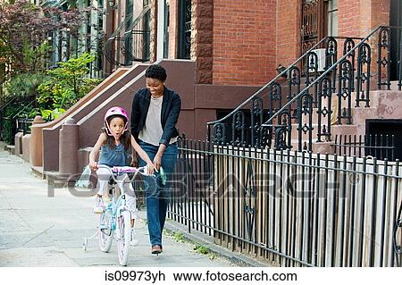 Daughter learning to ride bicycle along sidewalk with mother View Large Photo Image Stock Image - Daughter learning to ride bicycle along sidewalk with mother. Fotosearch