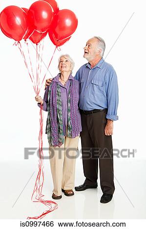 Stock Photograph - Senior couple holding red balloons against white background. Fotosearch