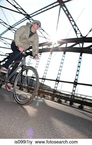 Stock Photo - Mid adult cyclist riding bike across bridge. Fotosearch