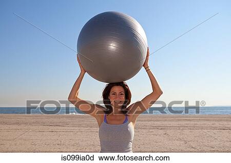 Stock Image - Woman on beach holding exercise ball. Fotosearch